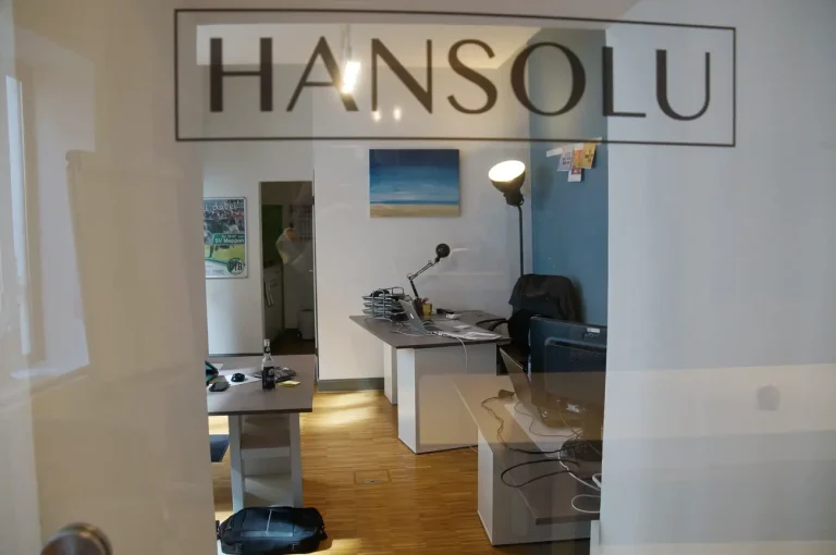 Unser Hansolu-Büro von innen. Man sieht durch eine Glastür mit Hansolu-Logo ein Büro mit drei Arbeitsplätzen.