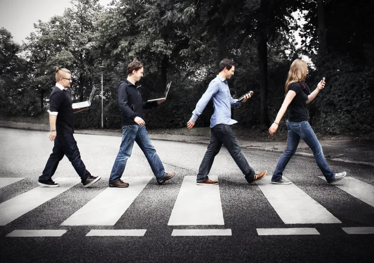 Fabi, Nils, Michi und Rebecca überqueren einen Zebrastreifen. Jeder schaut auf ein Gerät wie ein Laptop, Tablet oder Smartphone.
