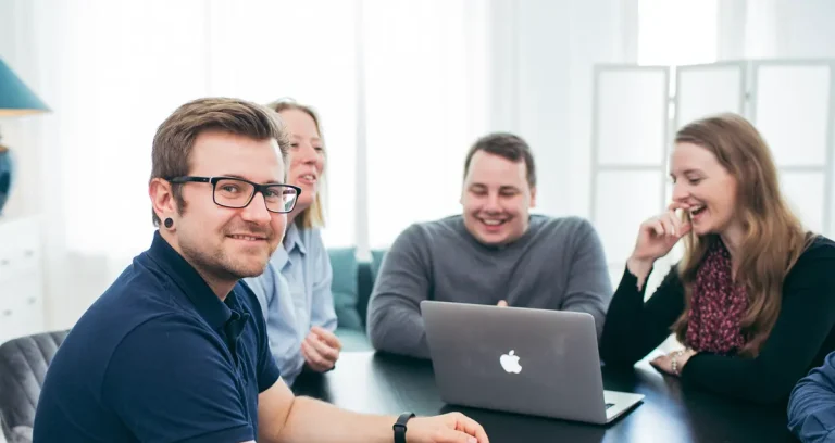 Fabian, Andrea, Paul und Rebecca sitzen am Meetingtisch. Fabian schaut lächelt in die Kamera und die anderen drei schauen auf eine Laptop und lachen.