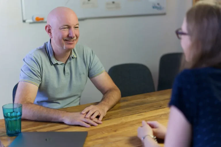 Unser Datenschutz-Experte Alex im Gespräch. Er sitzt am Meetingtisch und unterhält sich mit einer Frau.
