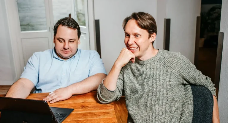 Nils und Paul im Gespräch. Sie sitzen am Meetingtisch und schauen auf einen Laptop.