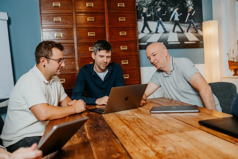 Alex, Michi und Fabian schauen auf einen Laptop. Sie sitzen im Meetingraum an einem Teakholztisch.