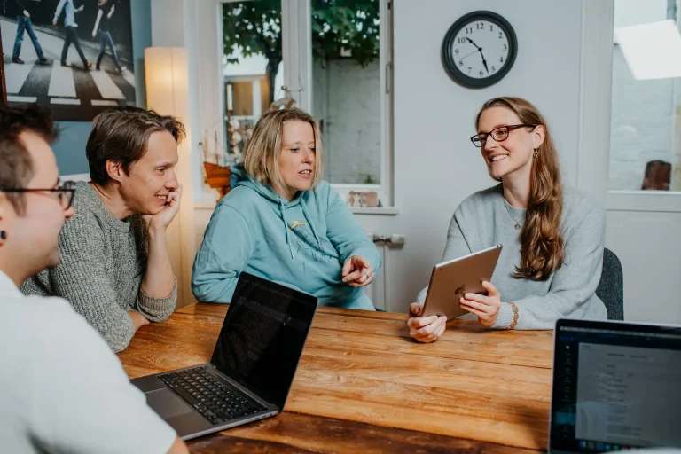 Rebecca zeigt Andrea, Nils und Fabi etwas auf einem Tablet. Sie sitzen in einem Meetingraum mit Teakholztisch.