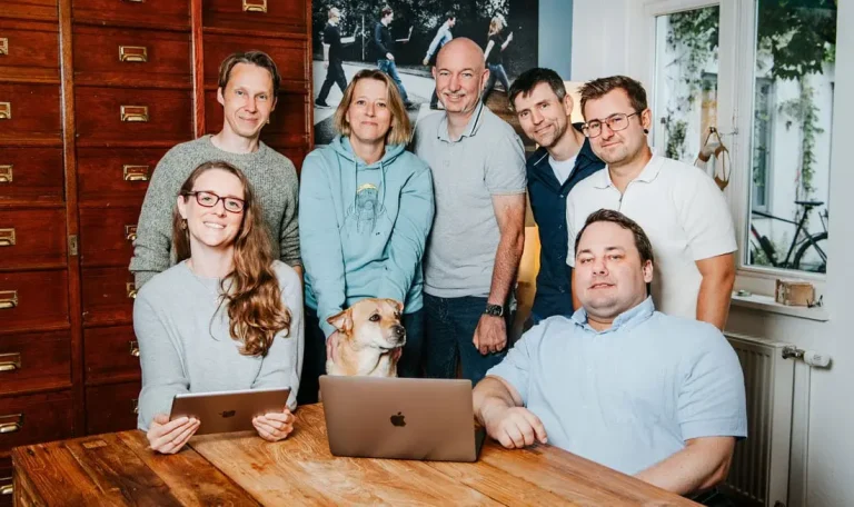 Teambild vom Hansolu-Team. Sie stehen im Meetingraum hinter einem Teakholztisch. Ein Laptop steht vor ihnen auf dem Tisch.