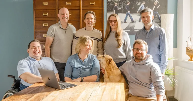Teambild vom Hansolu-Team. Sie stehen im Meetingraum hinter einem Teakholztisch. Ein Laptop steht vor ihnen auf dem Tisch.
