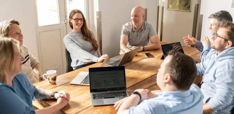 Das Hansolu-Team sitzt im Meetingraum an einem Holztisch und diskutiert. Mehrere haben einen Laptop vor sich.