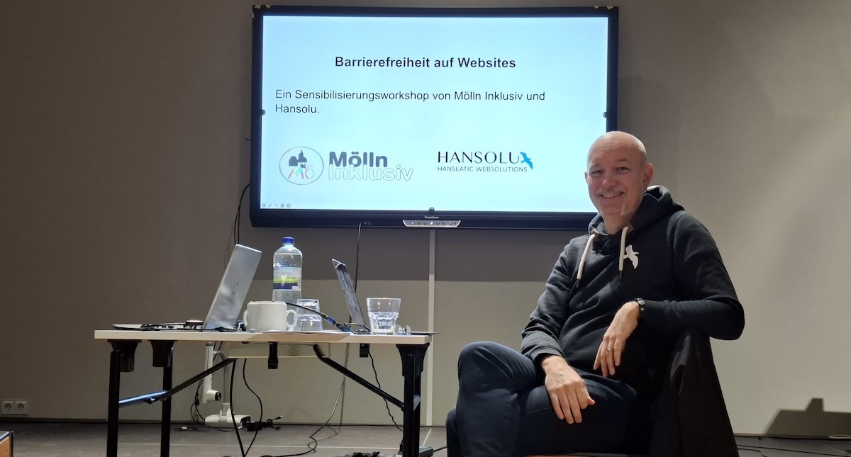 Barrierefreiheit Workshop: Alex sitzt auf einem Stuhl vor einem großen Monitor, auf dem der Titel des Workshops steht: Barrierefreiheit auf Websites.